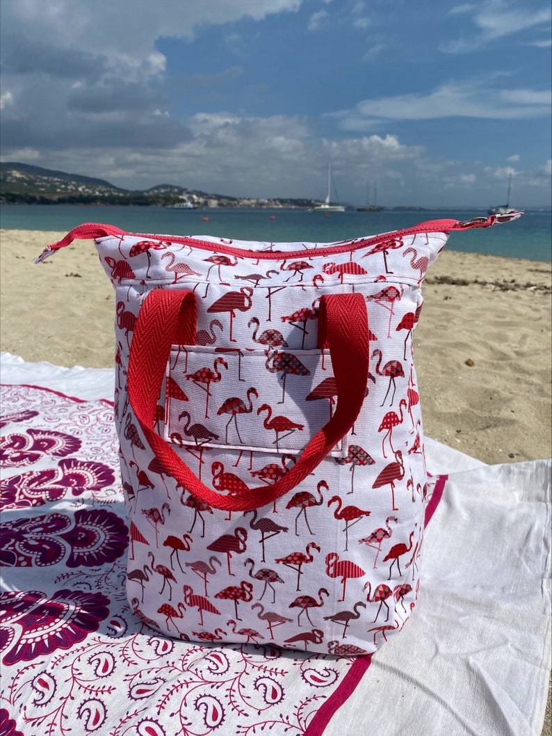 May include: A white tote bag with a red flamingo print and red handles. The bag is sitting on a white and pink patterned blanket.