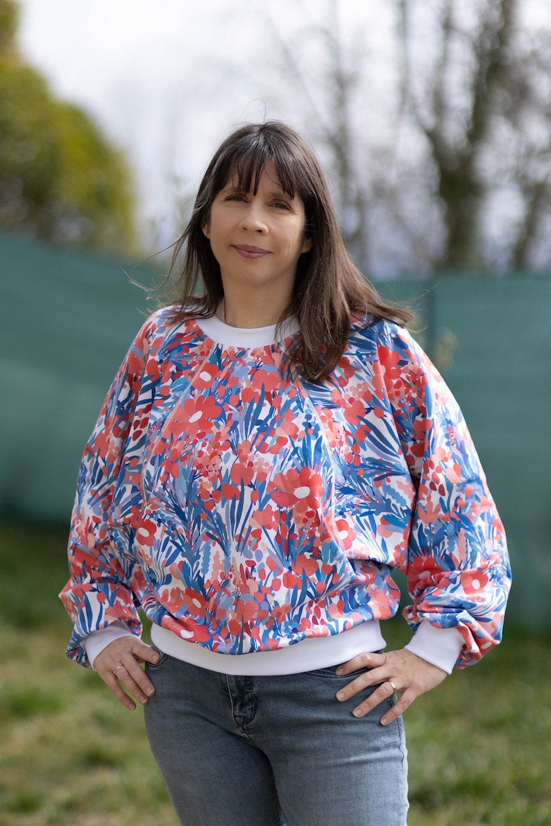 Pode incluir: Uma camisola branca com estampa floral em tons de vermelho, azul e rosa. A camisola tem um colarinho e punhos canelados brancos.