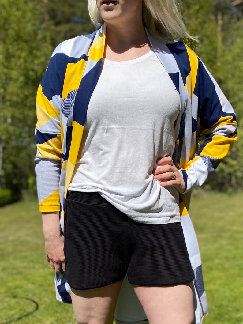 May include: A woman wearing a white t-shirt, black shorts, and a yellow, blue, and white patterned cardigan.