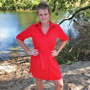 May include: A woman wearing a red button-down shirt dress with a belt, standing on a sandy beach near a body of water.