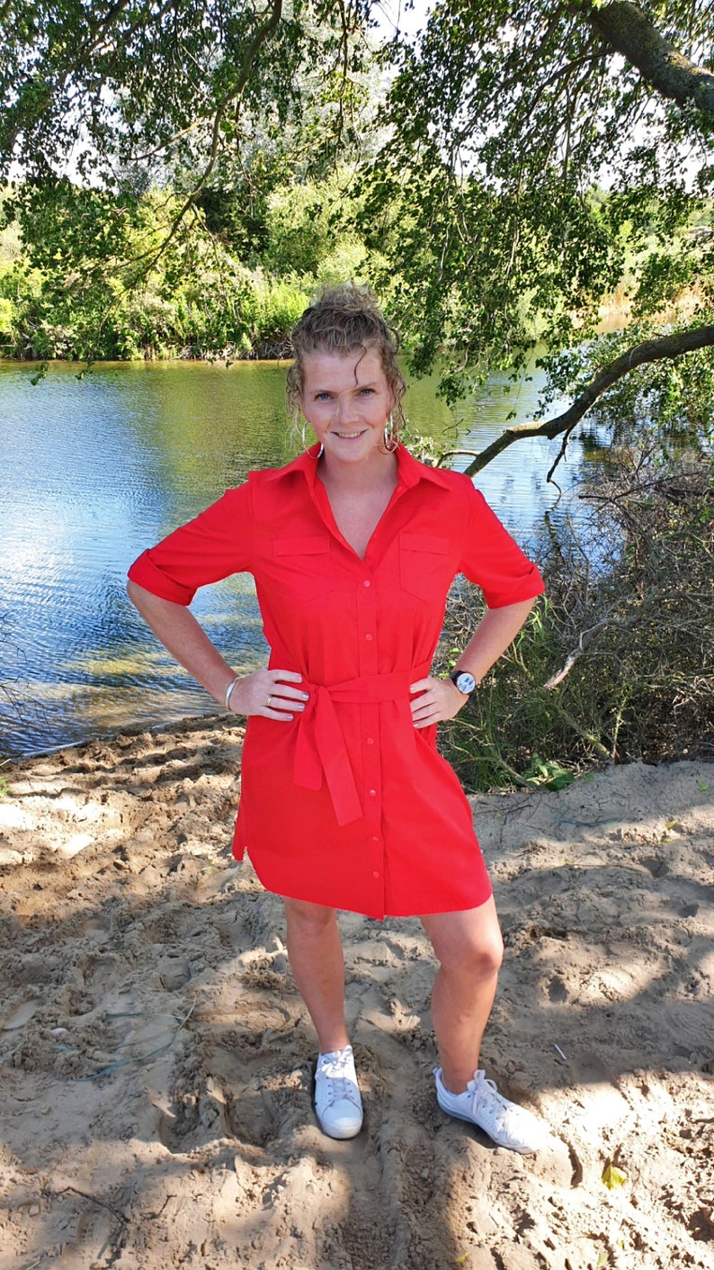 May include: A woman wearing a red button-down shirt dress with a belt, standing on a sandy beach near a body of water.