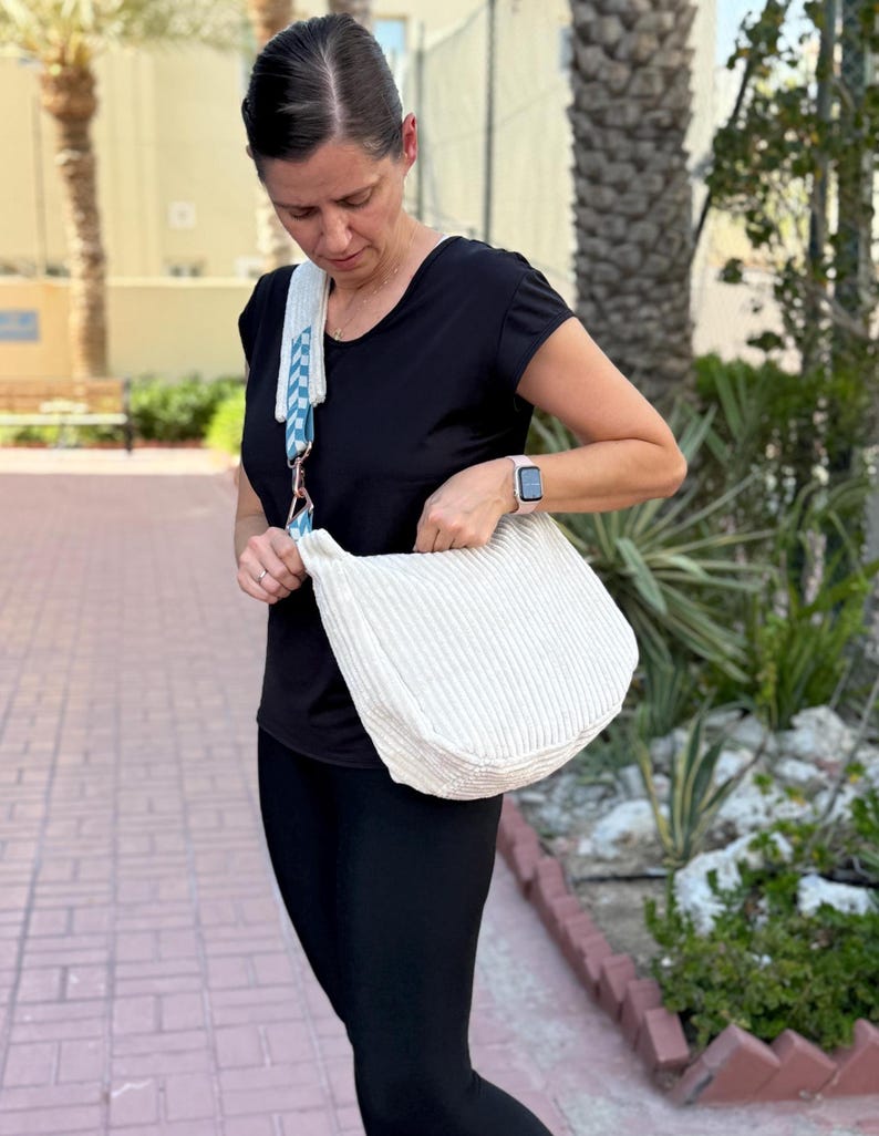 Puede incluir: Bolso de hombro a rayas color crema con una correa a cuadros azul y blanco. El bolso se lleva al hombro. La persona lleva una camiseta negra y leggings negros. El bolso parece estar hecho de una tela suave y texturizada.