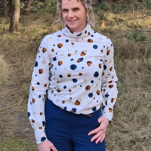 K&ouml;nnte beinhalten: Ein wei&szlig;es Rollkragen-Shirt mit einem braunen und blauen abstrakten Muster. Das Shirt hat lange &Auml;rmel und eine figurbetonte Silhouette. Die Person tr&auml;gt eine blaue Hose.