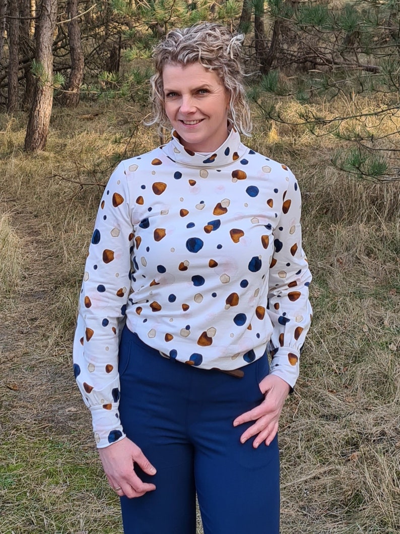 K&ouml;nnte beinhalten: Ein wei&szlig;es Rollkragen-Shirt mit einem braunen und blauen abstrakten Muster. Das Shirt hat lange &Auml;rmel und eine figurbetonte Silhouette. Die Person tr&auml;gt eine blaue Hose.