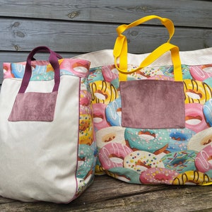 May include: Two canvas tote bags with donut patterns. The larger bag has a yellow handle and a pink and blue donut pattern. The smaller bag has a purple handle and a white background with a pink and yellow donut pattern. Both bags have a brown suede pocket on the front.