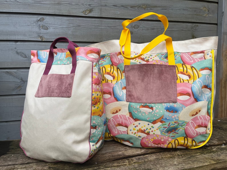 May include: Two canvas tote bags with donut patterns. The larger bag has a yellow handle and a pink and blue donut pattern. The smaller bag has a purple handle and a white background with a pink and yellow donut pattern. Both bags have a brown suede pocket on the front.