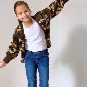 May include: A young person wearing a brown, green, and beige camouflage cardigan with a white t-shirt underneath. They are also wearing blue jeans.