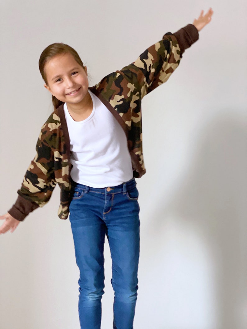 May include: A young person wearing a brown, green, and beige camouflage cardigan with a white t-shirt underneath. They are also wearing blue jeans.