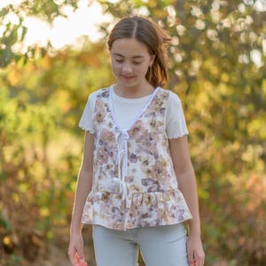 May include: A floral print vest with a white tie closure and a ruffled hem. The vest is layered over a white t-shirt and paired with light blue jeans. The background is a blurred view of a natural outdoor setting.