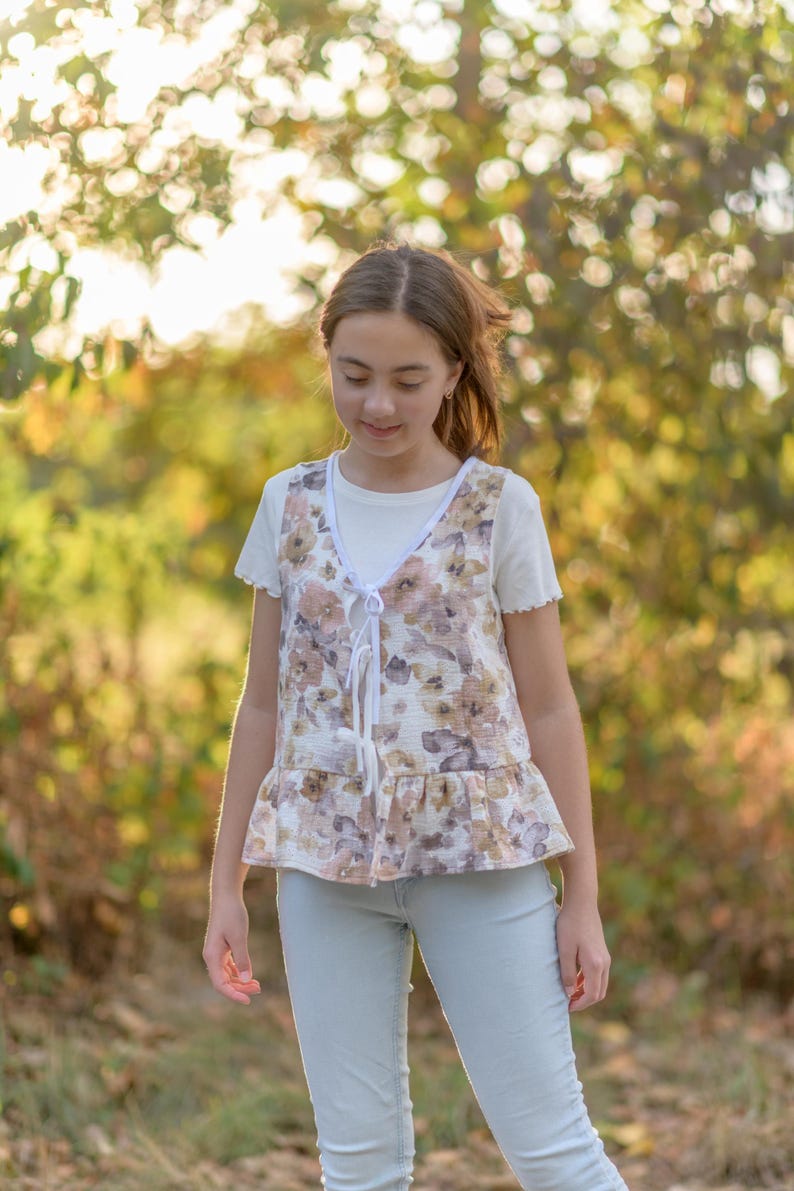 May include: A floral print vest with a white tie closure and a ruffled hem. The vest is layered over a white t-shirt and paired with light blue jeans. The background is a blurred view of a natural outdoor setting.