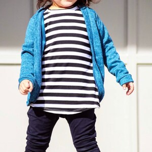 May include: A young child wearing a black and white striped shirt, a blue sweater, black pants, and white sneakers. The child is also wearing a black and white striped beanie.