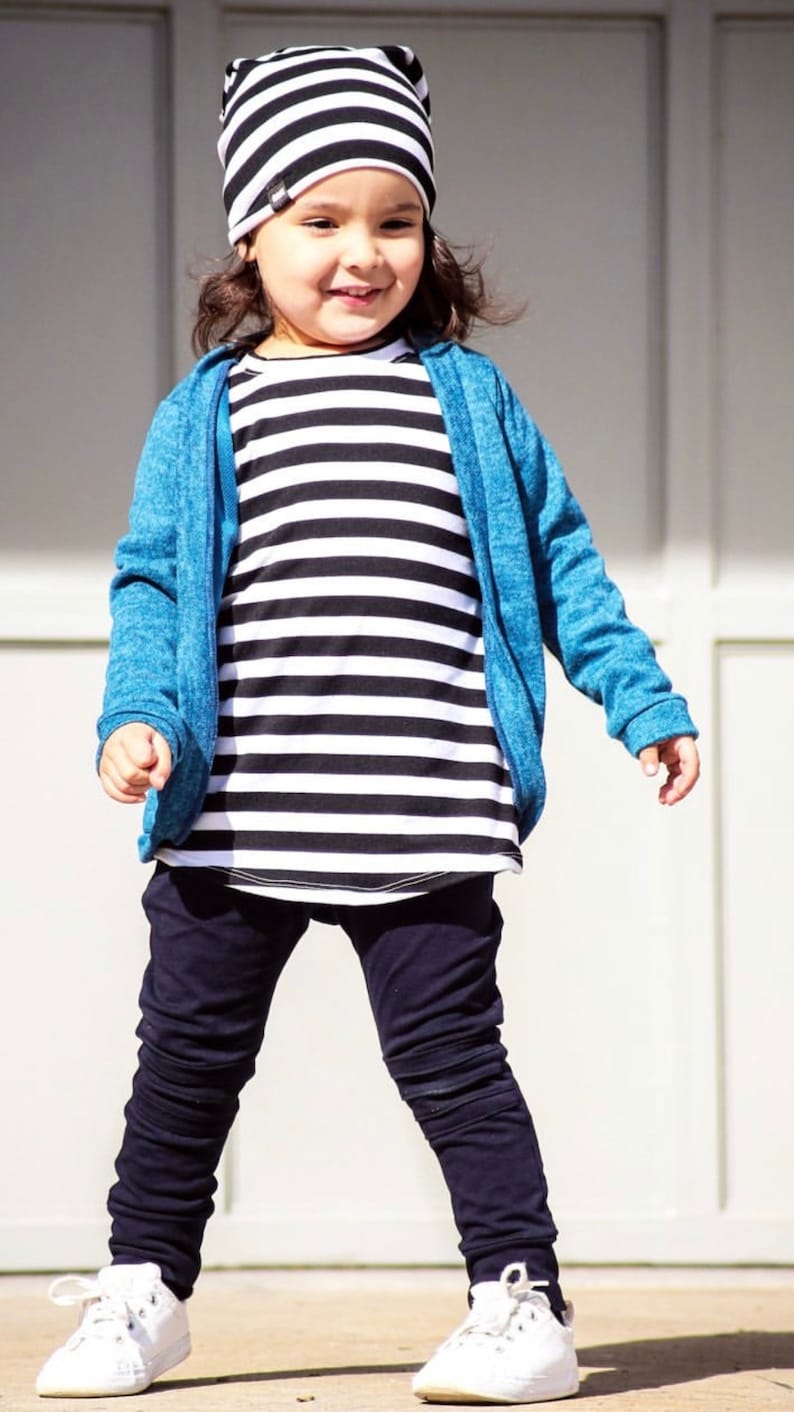 May include: A young child wearing a black and white striped shirt, a blue sweater, black pants, and white sneakers. The child is also wearing a black and white striped beanie.