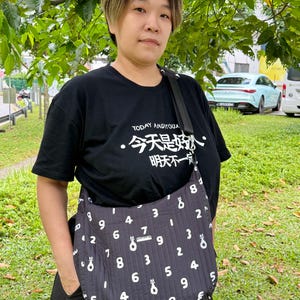 Puede incluir: Un bolso negro con un patr&oacute;n de n&uacute;meros blancos y una correa negra. El bolso se lleva cruzado. La persona lleva una camiseta negra con caracteres chinos y el texto "TODAY ANDIYOUX".
