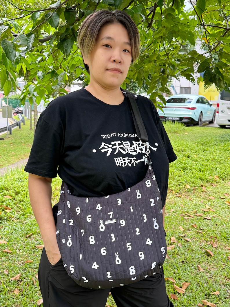 Puede incluir: Un bolso negro con un patr&oacute;n de n&uacute;meros blancos y una correa negra. El bolso se lleva cruzado. La persona lleva una camiseta negra con caracteres chinos y el texto "TODAY ANDIYOUX".