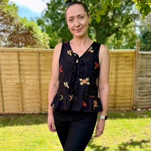 May include: A black sleeveless top with a V-neck and a tie closure. The top is adorned with colorful floral embroidery and a ruffled hem. The person is wearing black pants and sunglasses.