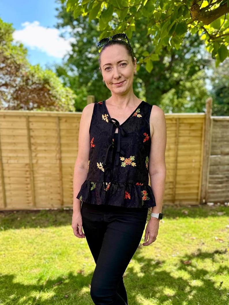 May include: A black sleeveless top with a V-neck and a tie closure. The top is adorned with colorful floral embroidery and a ruffled hem. The person is wearing black pants and sunglasses.