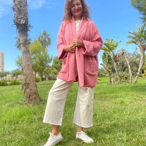 Pu&ograve; includere: Una donna indossa una giacca trapuntata rosa con una camicia bianca sotto e pantaloni bianchi larghi. &Egrave; in piedi in un parco erboso con palme sullo sfondo.