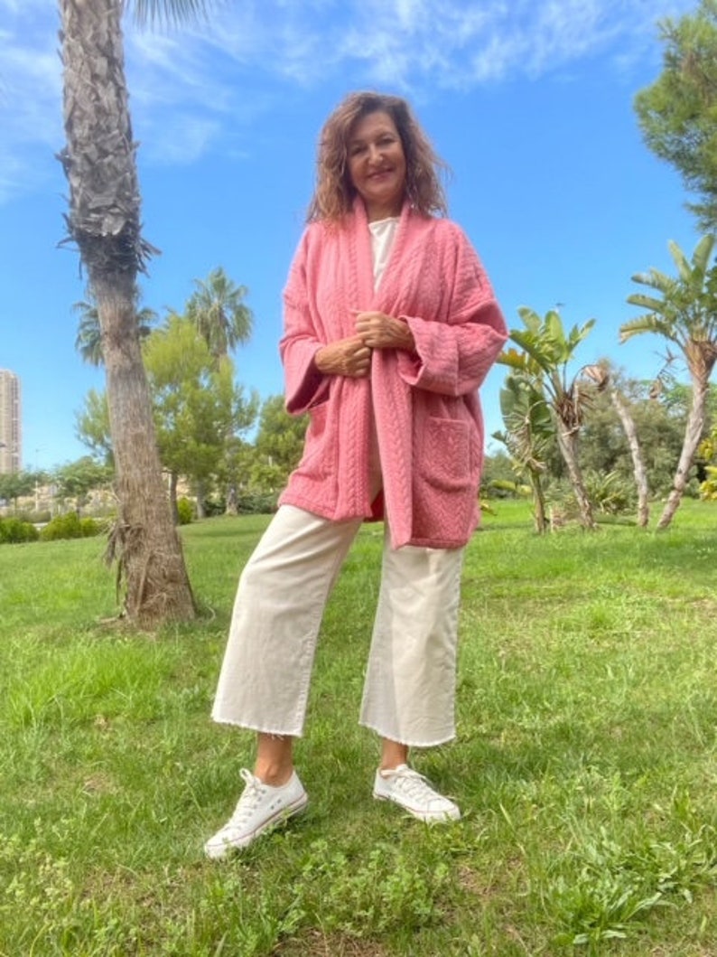 Pu&ograve; includere: Una donna indossa una giacca trapuntata rosa con una camicia bianca sotto e pantaloni bianchi larghi. &Egrave; in piedi in un parco erboso con palme sullo sfondo.