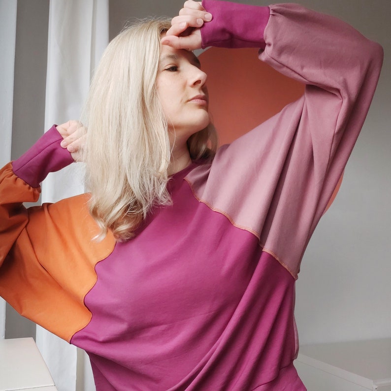 Pode incluir: Uma mulher vestindo um moletom rosa, laranja e roxo com blocos de cores, mangas compridas e um caimento solto.