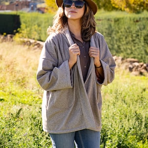 Pu&ograve; includere: Una donna con un cappello marrone, occhiali da sole, un cardigan grigio e jeans blu. &Egrave; in piedi in un campo di erba verde.