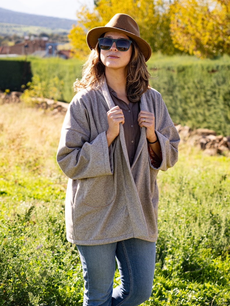 Pu&ograve; includere: Una donna con un cappello marrone, occhiali da sole, un cardigan grigio e jeans blu. &Egrave; in piedi in un campo di erba verde.