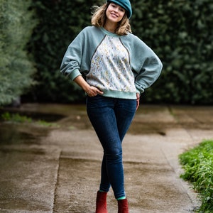 Pode incluir: Uma mulher vestindo um su&eacute;ter verde-azulado com um padr&atilde;o floral na frente, jeans azul e botas de tornozelo vermelhas. Ela est&aacute; usando um bon&eacute; de newsboy verde-azulado e sorrindo para a c&acirc;mera.