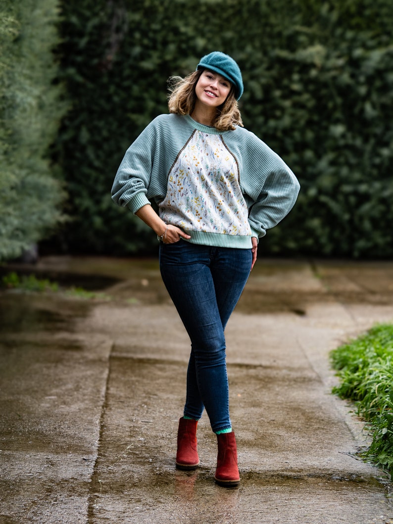 Pode incluir: Uma mulher vestindo um su&eacute;ter verde-azulado com um padr&atilde;o floral na frente, jeans azul e botas de tornozelo vermelhas. Ela est&aacute; usando um bon&eacute; de newsboy verde-azulado e sorrindo para a c&acirc;mera.