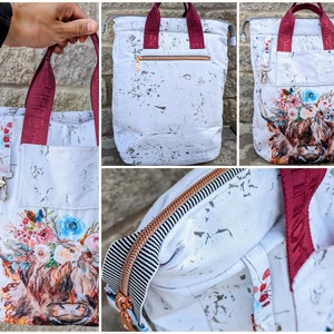 May include: White canvas tote bag with a watercolour highland cow design and floral accents. The bag has burgundy straps and a zipped pocket on the front. The bag is shown in three different views.