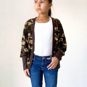May include: A child wearing a camouflage print cardigan with brown trim, a white tank top, and blue jeans.