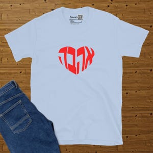 Light blue t-shirt spread on a brown parquet with blue jeans on left.  Design the Hebrew word ahava in red heart shape. Approx total design size 7 x 6.4 inches.