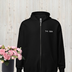 May include: Black zip-up hoodie with the Hebrew text "Eshet Chayil" embroidered on the upper chest. The hoodie has a drawstring hood and a full-length zipper. A bouquet of pink flowers sits to the left of the hoodie.
