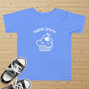 May include: A light blue t-shirt with a white cupcake graphic and the Hebrew text "Happy Birthday" on it.