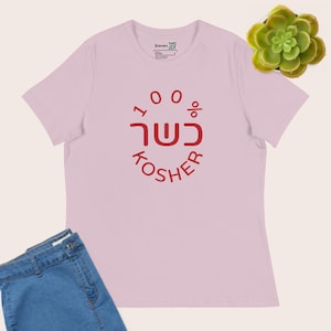 May include: A light pink short-sleeved t-shirt featuring a red circular graphic with the text 100% KOSHER and Hebrew characters in the centre. The shirt has a crew neckline and a small brand label printed on the inside of the collar area.