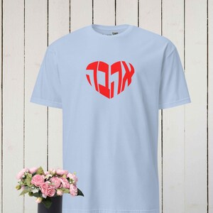 Light blue t-shirt, ghost mockup in front of beige wooden wall with pink roses. Design the word ahava in red Hebrew letters in a heart shape. Approx total design size 7" x 6.4".