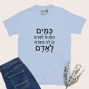 May include: A light blue t-shirt with black Hebrew text. The shirt has a crew neck and short sleeves. The text is arranged in four lines. The shirt is paired with blue jeans.