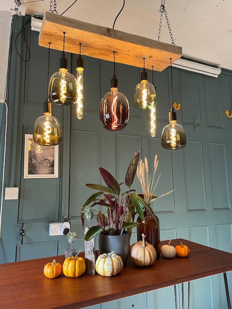 Może przedstawiać: Rustykalna drewniana lampa z sześcioma wiszącymi lampami Edison, niekt&oacute;re z przezroczystym kloszem szklanym, a inne z kolorowym kloszem szklanym. Lampa wisi nad stołem z dyniami i roślinami.