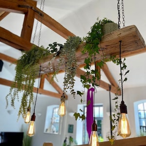 May include: A rustic wooden beam with hanging Edison bulb lights and greenery. The beam is suspended from the ceiling with chains and the bulbs are wired with black cords. The greenery is cascading down from the beam, adding a natural touch to the space.
