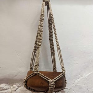 May include: A hanging macrame plant holder with a brown wooden pot. The macrame is made of tan-coloured rope, and the pot is rectangular. The plant holder is suspended from a single rope.