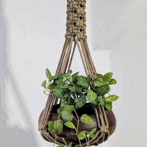 Può includere: Un portavaso in macramè color beige naturale, che sostiene un vaso di legno marrone scuro pieno di piante verdi con venature bianche. Il portavaso è sospeso da un anello, con il vaso assicurato da corde annodate.