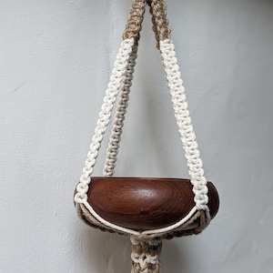 Könnte beinhalten: Ein Makramee-Pflanzenhänger mit einem braunen Holztopf. Der Hänger ist aus Naturkordel gefertigt und hat ein weiß-braunes Farbschema.