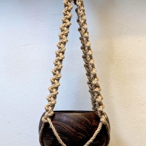 Puede incluir: Un macetero colgante de macramé hecho con cuerda de yute natural, con una maceta de madera marrón oscuro. El macetero está suspendido de un anillo de madera.