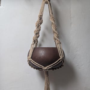 May include: A dark brown wooden bowl held by a beige macrame plant hanger. The hanger features twisted cord sections, a central knot supporting the bowl, and a long tassel extending from the bottom. A circular metal ring sits at the top.