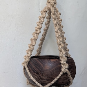 May include: A dark brown wooden bowl held by a beige macrame plant hanger made of thick twisted rope. The hanger features intricate knotted patterns that secure the bowl, which has a visible wood grain, and it hangs from a circular wooden ring.