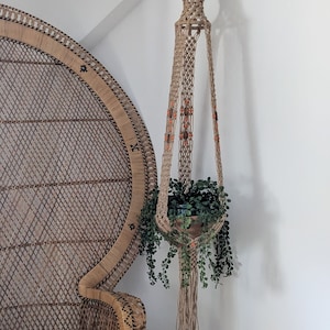 Pode incluir: Um suporte de plantas em macramé com uma planta em vaso, suspenso de um gancho de madeira numa parede branca. O suporte é adornado com contas laranja. Uma cadeira de vime em forma de pavão é parcialmente visível no fundo. Mede cerca de 100 cm.
