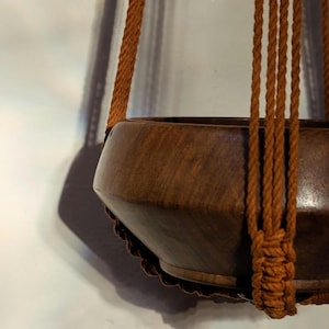 May include: A brown wooden hanging planter with a scalloped edge. The planter is suspended by thick, woven, orange-brown rope. The planter is empty and ready for a houseplant. The background is a light gray wall.