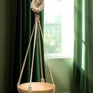 May include: A hanging wooden planter suspended by natural rope. The planter is a light brown bowl shape, held by a woven rope design. The rope is knotted at the top and hangs in front of a window with green curtains.