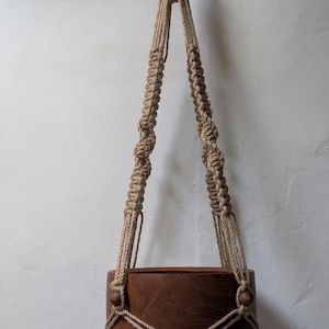 Pode incluir: Um suporte de plantas de macramé com um vaso de madeira marrom natural. O suporte é feito de corda natural e tem um design simples.