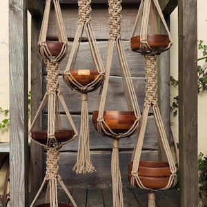 Op de afbeelding: Een verzameling macramé plantenhangers met houten kommen. De hangers zijn gemaakt van bruin touw en hangen aan een houten frame. De houten kommen hebben een rijke, donkerbruine kleur, wat een natuurlijke, bohemien uitstraling creëert.