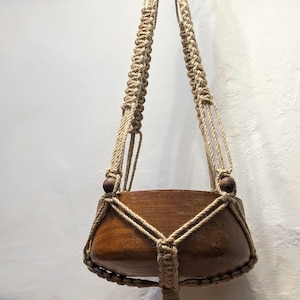 Puede incluir: Un macetero colgante de macramé con una maceta de madera. El macetero está hecho de cuerda de color natural y tiene cuentas de madera en la parte superior. La maceta es marrón y tiene una forma redondeada.