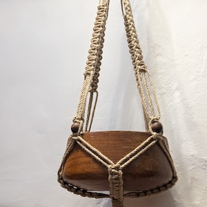 May include: A macrame plant hanger with a wooden pot. The hanger is made of natural beige cord and has wooden beads at the top. The pot is brown and has a rounded shape.